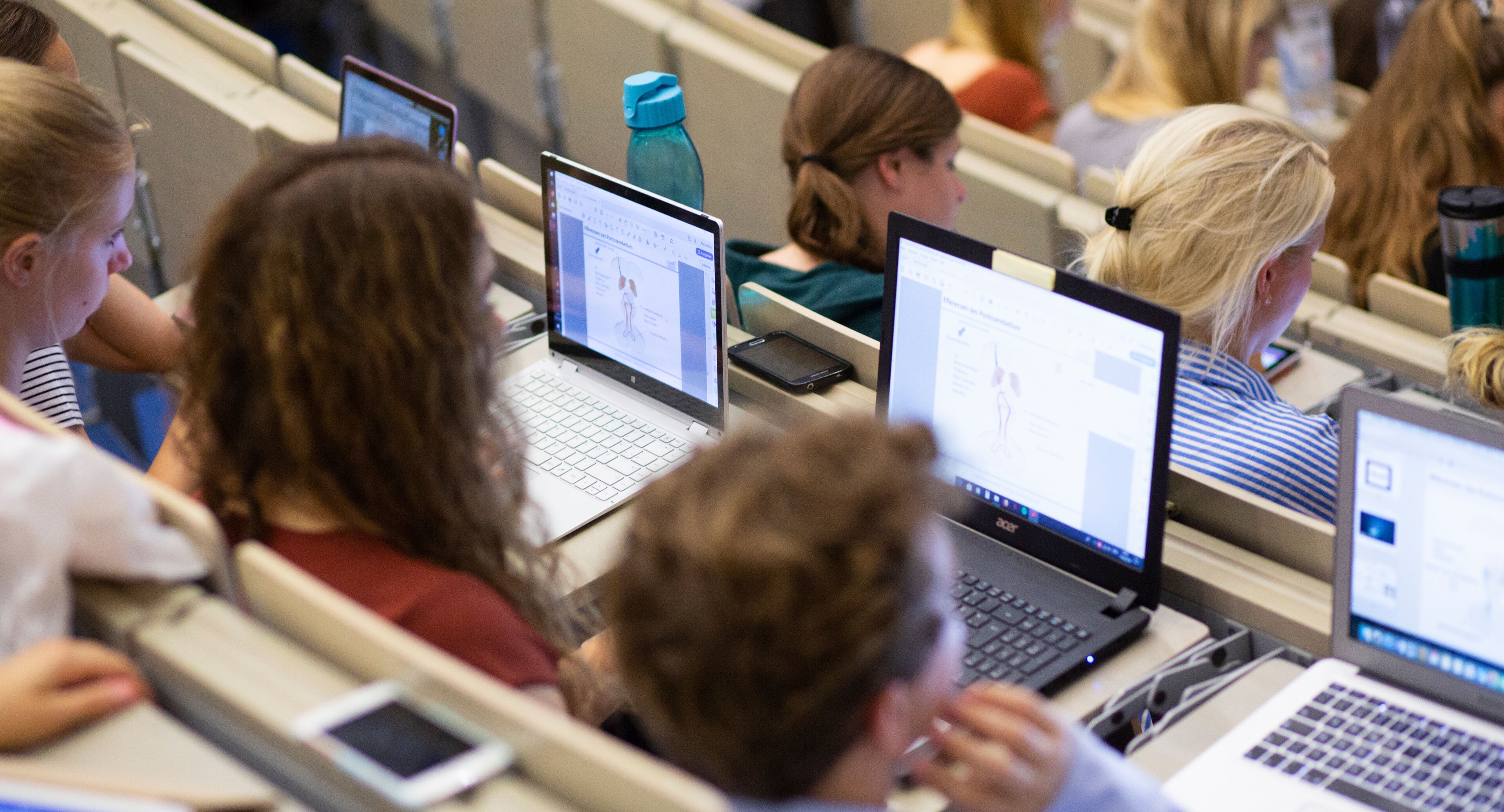 In dem Bild sind mehrere Nutzer zu sehen, die in einem Vorlesungsraum in Reihen sitzen, die von vorne nach hinten in die Höhe gehen. Die Nutzer haben aufgeklappte Laptops oder Tablets vor sich liegen.